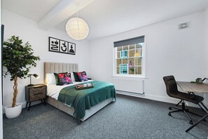 A bedroom with a bed, bedside table, lamp, potted plant, and a window with a view of a building.