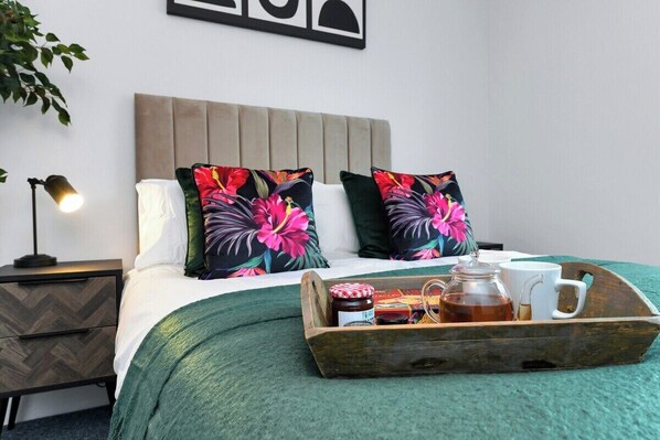 A neatly made bed with floral pillows, a bedside lamp, a wooden tray with a teapot and cups, and a herringbone-patterned nightstand.