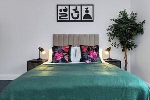 A bed with a teal bedspread, two nightstands with lamps, a wall-mounted artwork, and a potted plant.