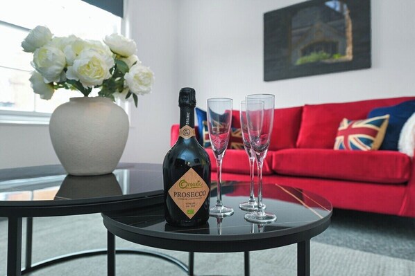 A bottle of Organic Prosecco on a glass table with two empty wine glasses, a vase of white flowers, and a red sofa with a Union Jack pillow in a modern living room.