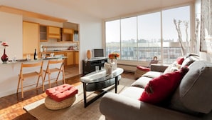 En moderne stue med sofa, skammel og sofabord, et køkkenområde med træskabe, og et stort vindue med udsigt over byen.