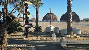 A beach with a wooden deck, palm trees, and a signpost with various destination names. - Luxury apartment -  Sea, Mountain, Golf - La Duquesa Manilva (Manilva)