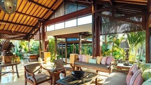 A spacious indoor area with a high ceiling, wooden furniture, and large windows offering views of greenery outside. - Villa Semarapura | 5BR Luxury Oceanfront Villa | Pool & Full Staff (Kabupaten Badung)