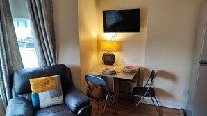 Un salon avec un canapé en cuir noir, une petite table à manger avec des chaises, un téléviseur à écran plat fixé au mur et une lampe sur la table.