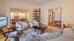 A modern living room with a sofa, armchair, coffee table, and bookshelf. There is a dining area with a table and chairs in the background.