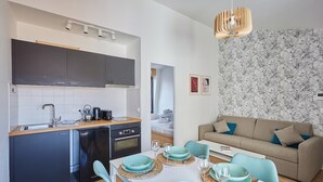 Een compacte keuken met moderne apparaten, een eetgedeelte met blauwgroene stoelen en borden, en een woonkamer met een bank en behang met een patroon.