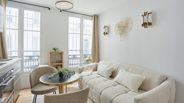 A modern living room with a beige sofa, a round table with chairs, a bookshelf, and a balcony with a view.