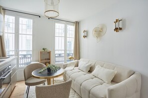 Una sala de estar moderna con un sofá color crema, una mesa redonda con sillas, una estantería y un balcón con vistas.