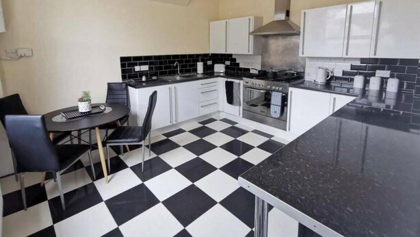 A kitchen with a black and white checkered floor, white cabinets, a stove, and a dining area with a table and chairs. - 5 Bedroom Holiday House – Clarendon 65 (Wallasey)