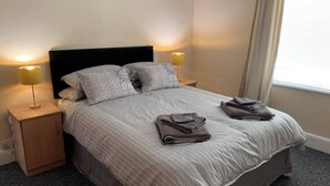 A neatly made bed with two pillows, a bedside lamp, and folded towels on a carpeted floor. - 5 Bedroom Holiday House – Clarendon 65 (Wallasey)