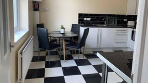 A compact kitchen with a checkered floor, a dining table with chairs, and white cabinetry. - 5 Bedroom Holiday House – Clarendon 65 (Wallasey)