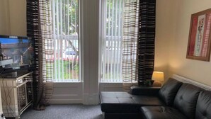 A living room with a sectional sofa, a coffee table, and a TV on a stand. There are curtains on the windows, and a lamp on a side table. - 5 Bedroom Holiday House – Clarendon 65 (Wallasey)
