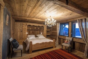 A wooden cabin bedroom with a bed, a chair, a chandelier, and a window with curtains.