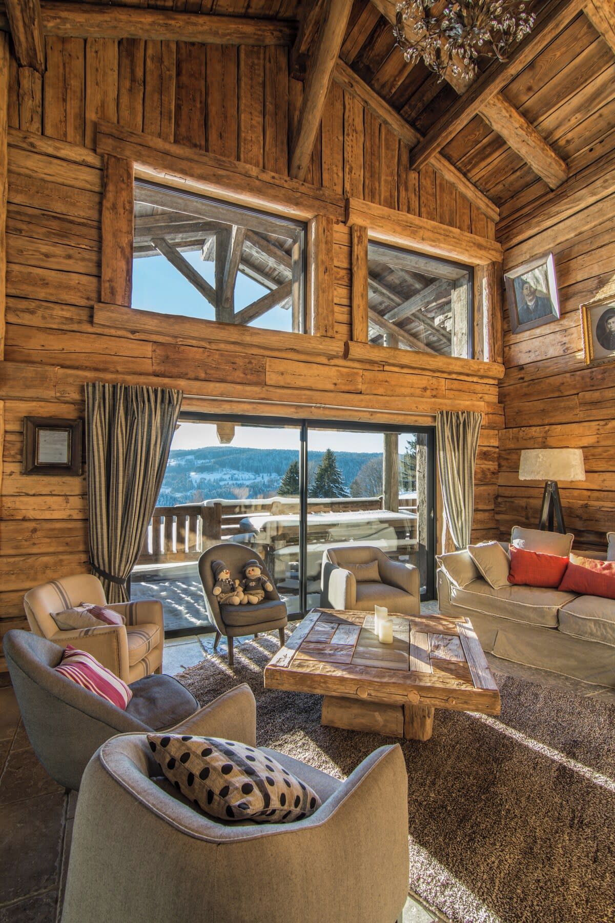 A log cabin interior with a large window, furnished with leather sofas and armchairs, a wooden coffee table, and a view of a snowy landscape.