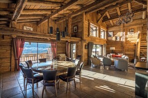 A rustic wooden interior with a dining area, seating, and a view through large windows.