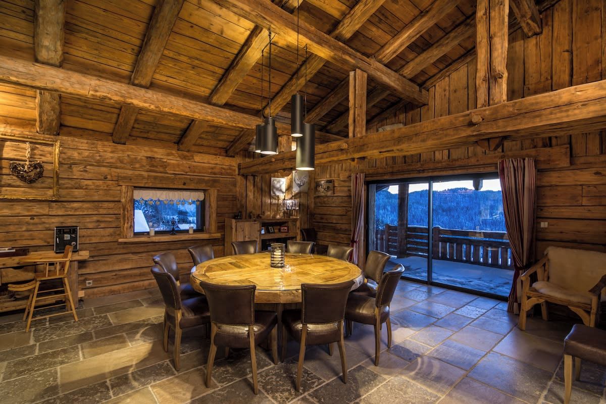 A wooden interior with a round table and chairs, a fireplace, and a large window with a view.
