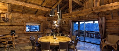 A wooden interior with a round table and chairs, a fireplace, and a large window with a view.