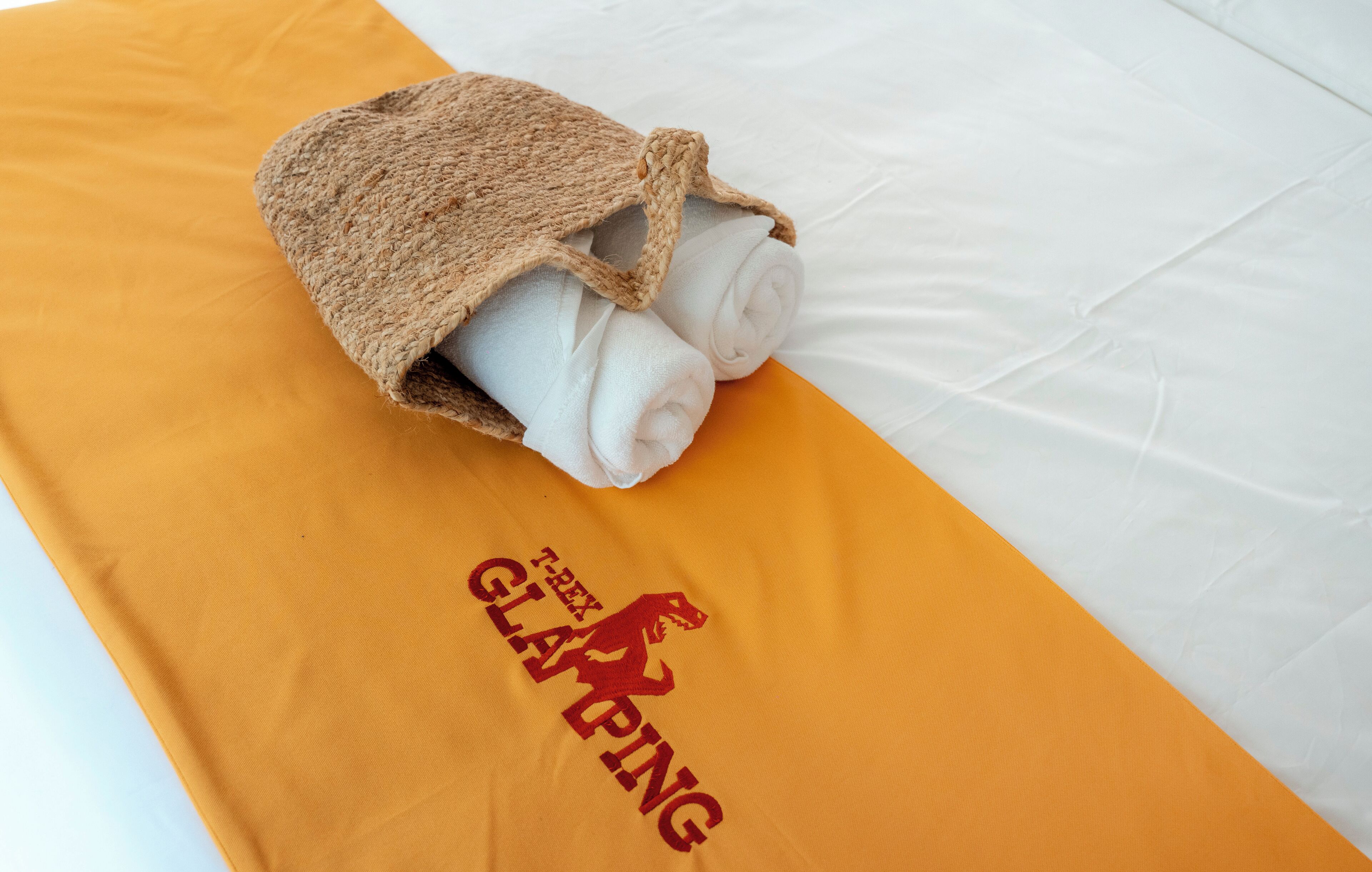 A bed with a rolled white towel and a brown head covering on an orange towel with a logo.