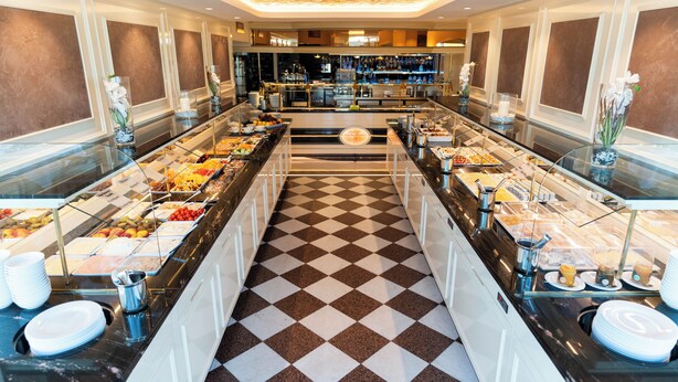 Uma área de buffet com piso xadrez, vitrines de vidro com comida e um bar ao fundo.