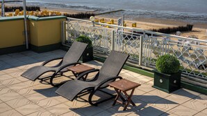 Terraço com duas poltronas de descanso e uma mesa pequena, com vista para uma praia com grade e torre salva-vidas.