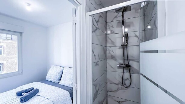 A modern bathroom with a glass shower enclosure, a bed with a blue patterned bedspread, and a window with a view of a building.