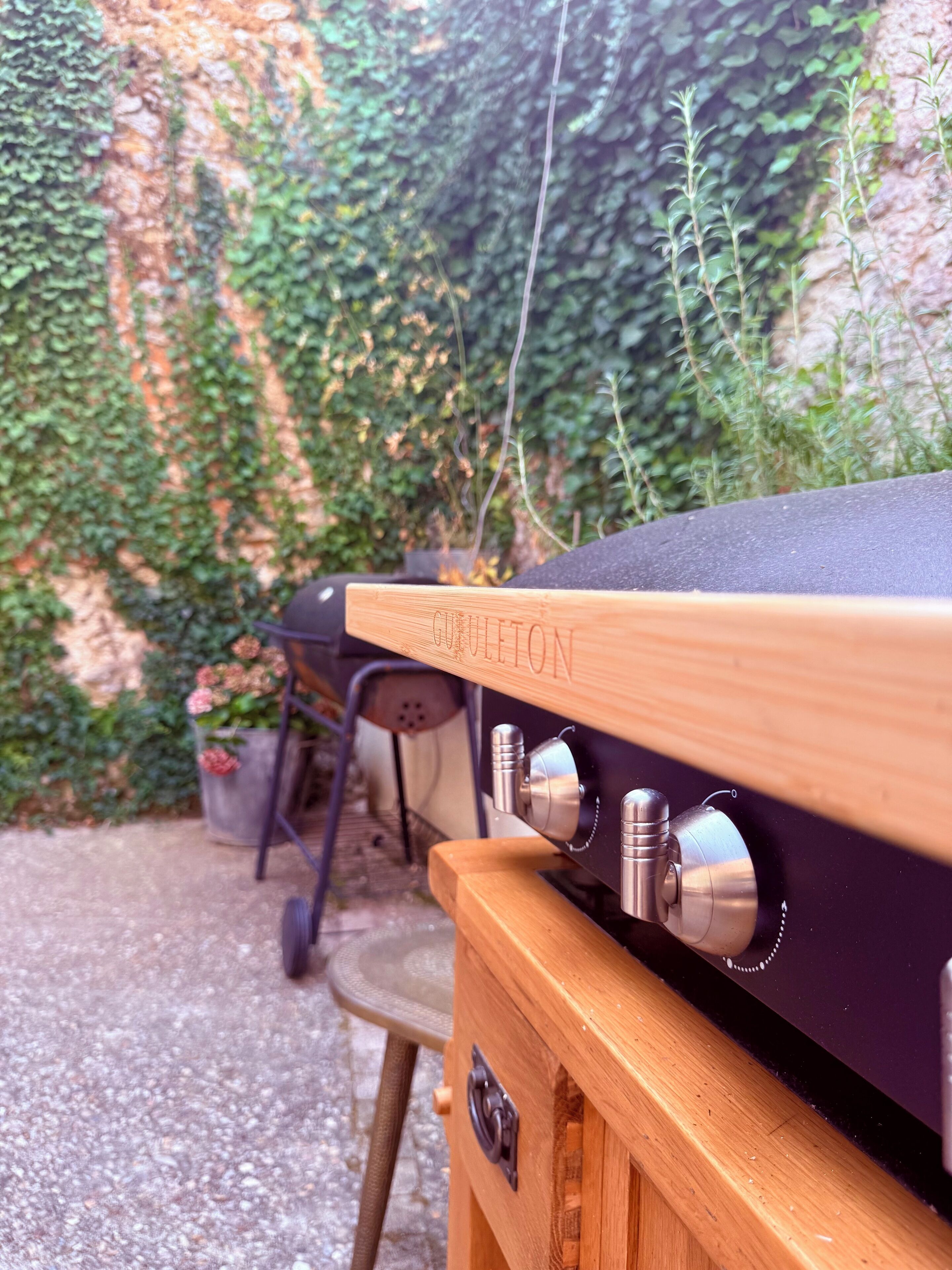 A barbecue grill with a wooden handle and a metal lid, set against a wall with ivy.