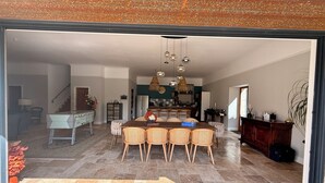 A spacious dining area with a large wooden table and wicker chairs, a kitchen in the background, and a staircase leading upstairs. - Le Patio Bleu (Saint-Romain)