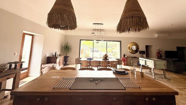 A spacious kitchen with a large island, two hanging woven light fixtures, a dining area with chairs, and a view through a large window. - Le Patio Bleu (Saint-Romain)