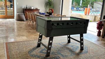 Amplo espaço interno com mesa de pebolim, mesa de jantar com cadeiras e vista para área externa com piscina.