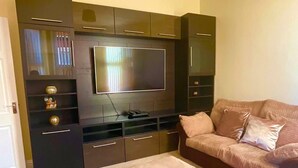 Een woonkamer met een flatscreen televisie, donkere kasten, een bank en een poef.