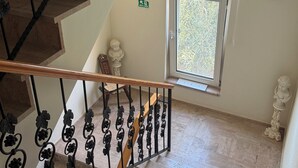 Eine Treppe mit hölzernem Geländer und schwarzen Metallreling mit floralen Verzierungen, die zu einer Zwischenebene mit einem Fenster und einer Statue führt.