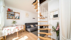 A cozy living room with a wooden staircase, a grey sofa, a wooden table with a floral tablecloth, and a bookshelf filled with books and decorative items. - Cute duplex studio, in the mountains, sleeps 5 (Allos)