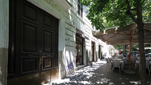 Una acera con mesas al aire libre, un edificio con puerta cerrada y un árbol que da sombra.