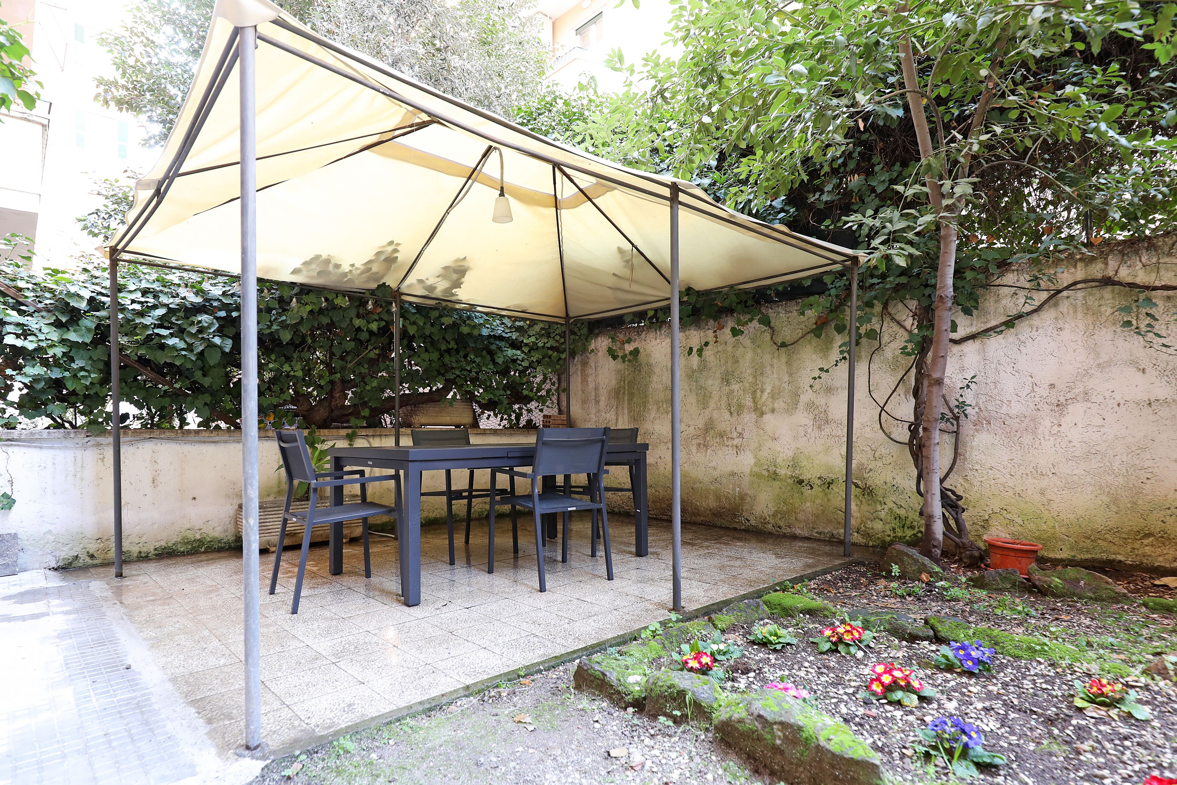 A covered patio with a table and chairs, surrounded by greenery and a garden.