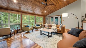 Un vaste salon doté de plafonds en bois, de grandes fenêtres offrant une vue sur la forêt et d’un coin salon confortable avec des canapés et des fauteuils.