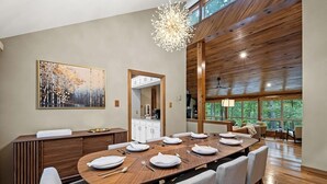 Un espace de salle à manger moderne, avec une table en bois dressée pour six personnes, une grande peinture abstraite accrochée au mur et une vue sur la forêt par de grandes fenêtres.