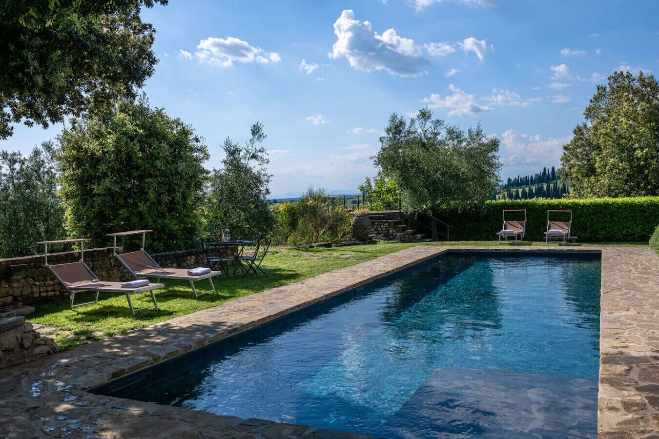A rectangular swimming pool with a stone border, surrounded by a grassy area and trees. There are lounge chairs and a table with chairs nearby.