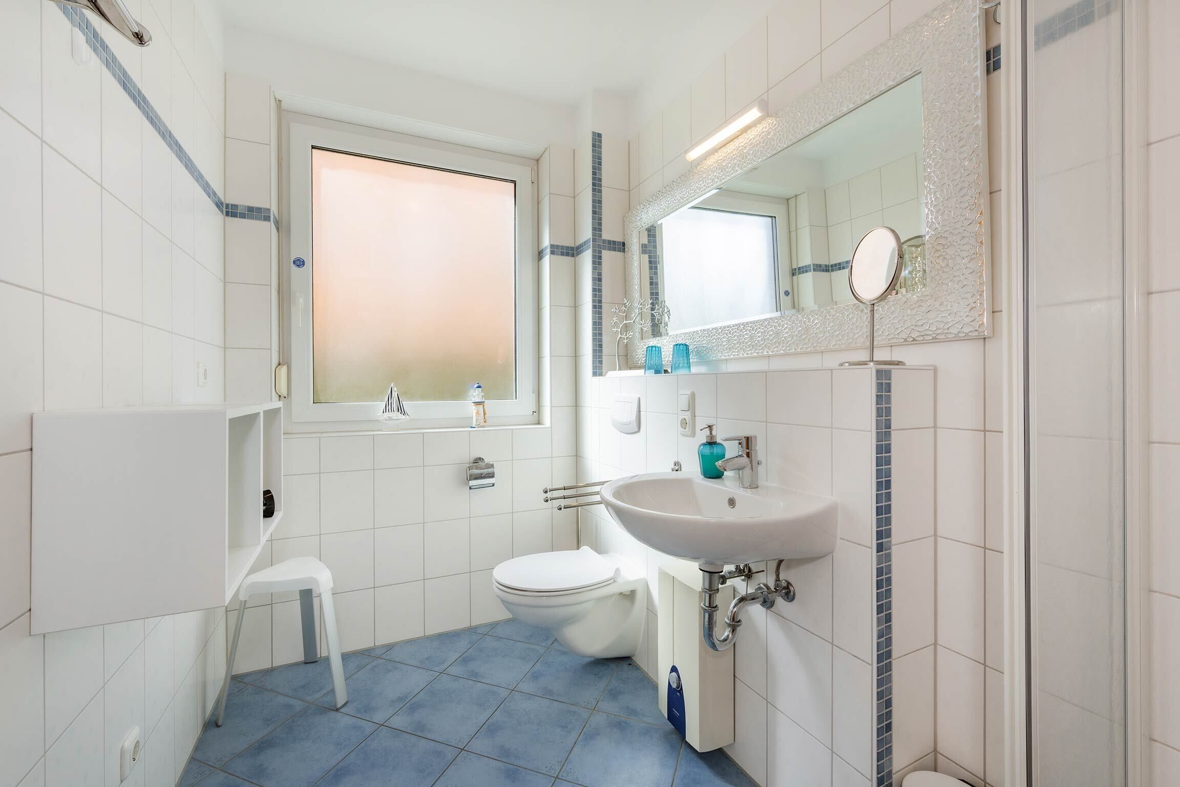 A bathroom with a white sink, a toilet, a small white stool, a window, and a mirror with a decorative frame.