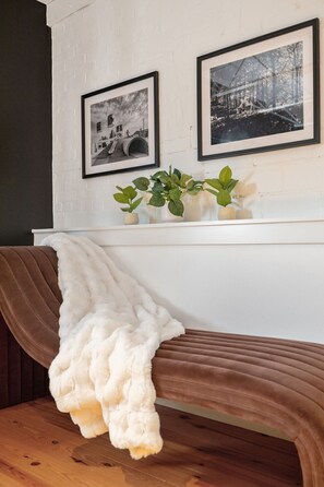 Una habitación con pared de ladrillo blanco, dos fotografías blanco y negro enmarcadas, un estante blanco con plantas verdes y un banco marrón con una manta blanca.