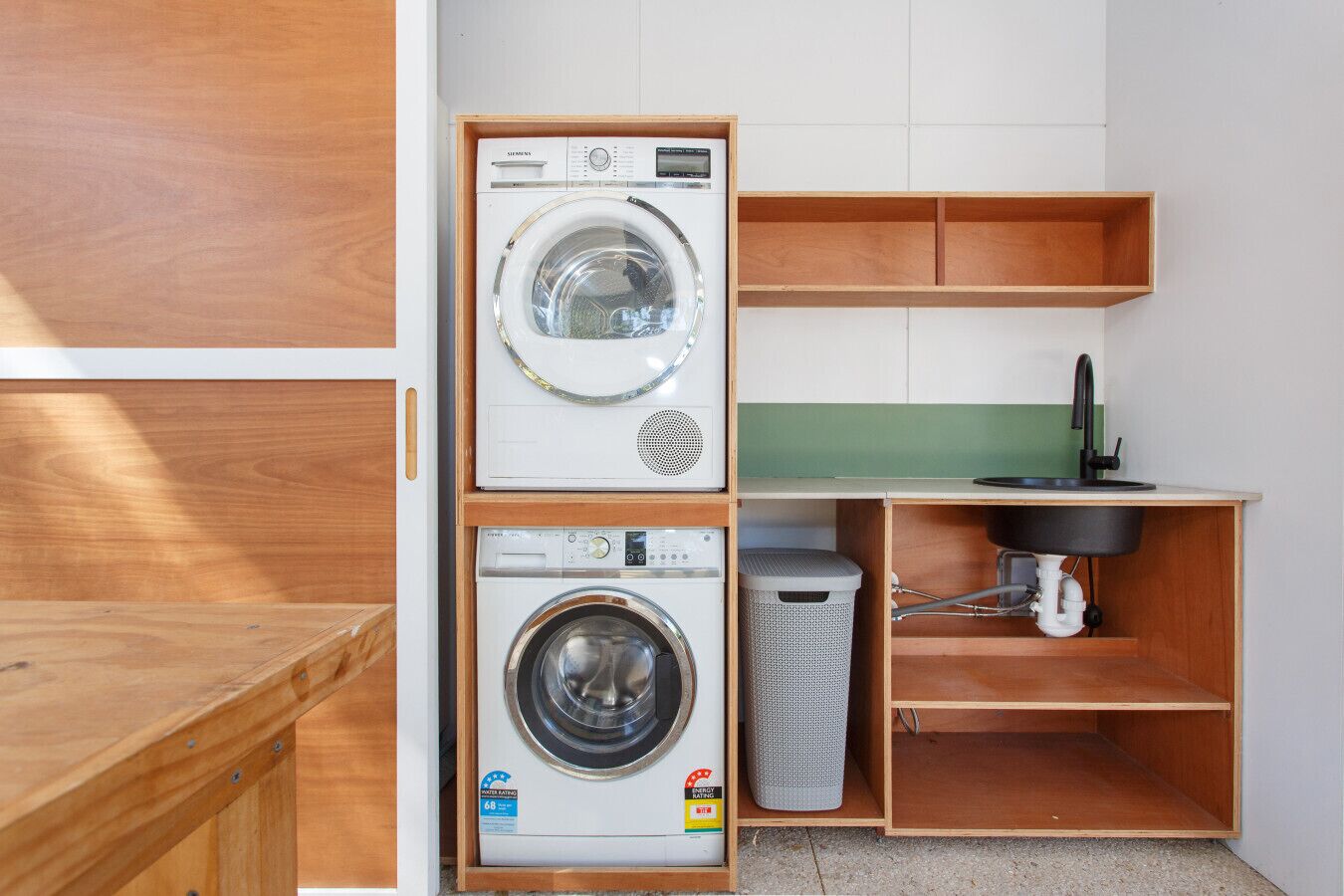 A compact laundry room with two stacked washing machines, a sink, and a trash bin.