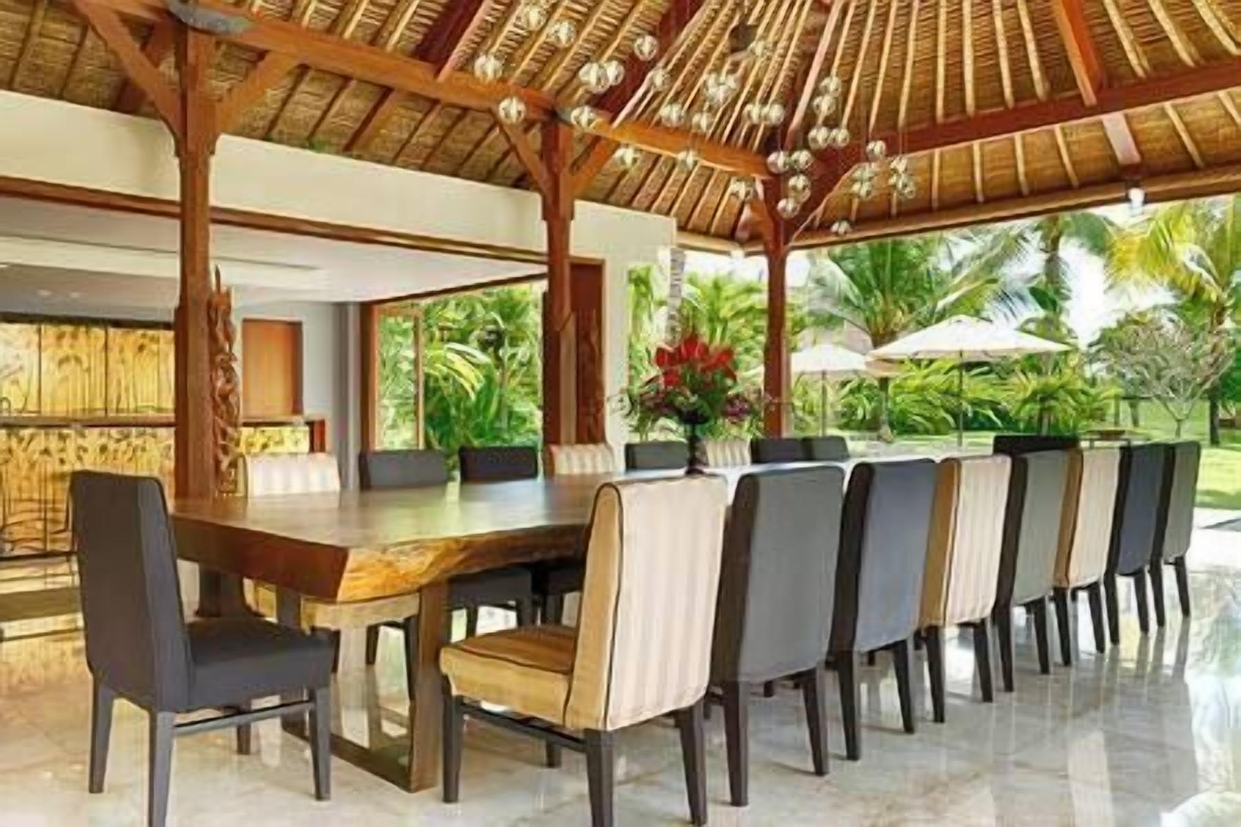Un'ampia zona pranzo all'aperto con tavolo e sedie in legno, tetto di paglia e vista sulla vegetazione tropicale.