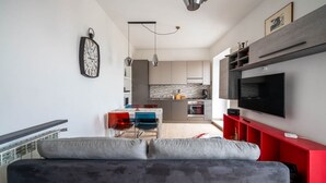A modern living room with a grey sofa, a red entertainment unit, and a kitchen in the background. - Al Belvedere A – Panoramic apartment in the Castelli Romani (Rocca di Papa) (Rocca di Papa)
