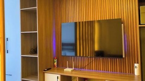 A room with a large flat-screen TV mounted on a wooden wall, a white cabinet to the left, and a small table with a remote control and a decorative item. - Smile's Home (Nairobi)