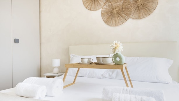 Una cama bien hecha con sábanas blancas, una mesita de noche con una lámpara y una bandeja con tazas y una flor.