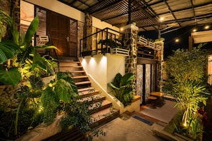 Un escalier avec un mur en pierre et des plantes, menant à un espace extérieur couvert, aménagé avec des tables et des chaises.