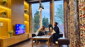 Una habitación con televisión, sofá, mesa y ventana con cortinas.