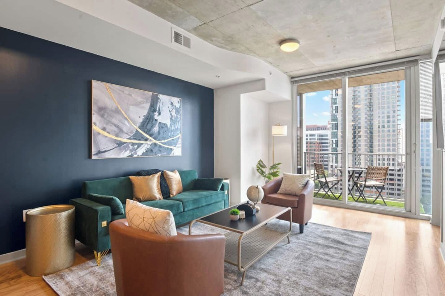 A modern living room with a green sofa, a brown armchair, and a coffee table. There is a large window with a view of high-rise buildings.