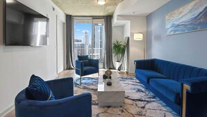 A modern living room with a large TV, two blue sofas, a coffee table, and a view of the city skyline through a large window.