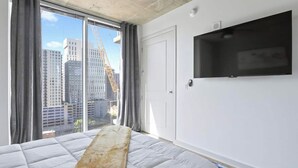 A bedroom with a large bed, a TV mounted on the wall, and a view of tall buildings outside.