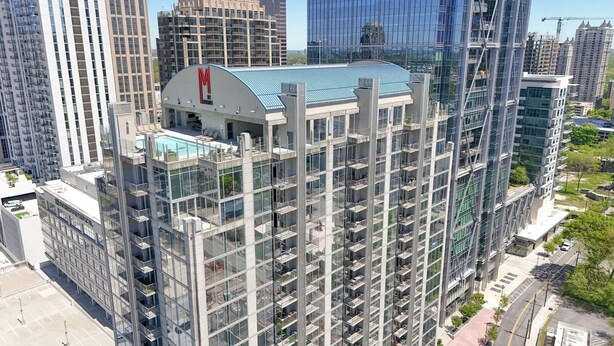 A high-rise building with a prominent 'M' logo, multiple balconies, and a rooftop terrace.